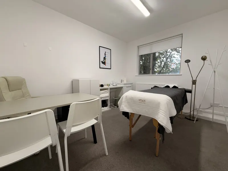 Sala de tratamiento con una camilla cubierta con una toalla de Vitalis, un escritorio con sillas y una ventana con vistas a los árboles